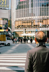 A street photography print of an elderly man in a checkered scarf waiting to cross the zebra stripes of Shibuya Crossing in Tokyo, with a white mini-truck and modern architecture in the background.