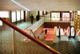A 35mm film photograph of businessmen walking through the mid-century modern corridors of the original Hotel Okura in Tokyo, featuring geometric patterned carpets and architectural staircases.