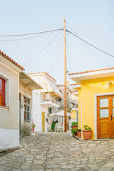 A simple photo of a tall wooden pole with wires connected to white houses under a bright blue sky in a Greek village.
